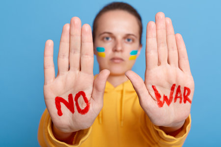 Serious patriotic woman in yellow hoodie with Ukrainian flag on cheeks, showing her palms with no war inscription, posing isolated over blue background.の写真素材