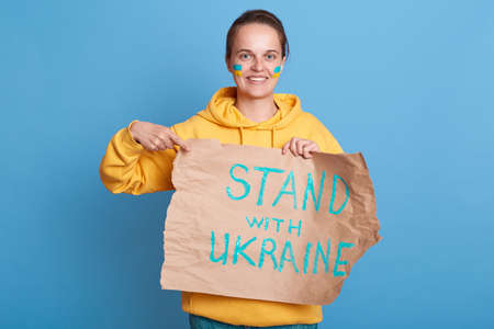 Smiling delighted woman in yellow hoodie with Ukrainian flag on cheeks, holding pointing placard with stand with Ukraine inscription, posing isolated over blue background.の写真素材
