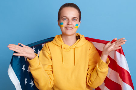 Pleasant kind woman wearing yellow hoodie with Ukrainian flag on cheeks, spreading hands, welcome, holding USA flag, looking at camera, posing isolated over blue background.の写真素材
