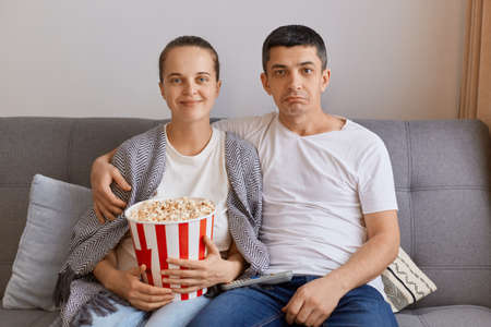 Indoor shot of young couple watching TV at home, sitting on cough, hugging, wife wrapped in plaid and holding bucket with popcorn, family spending time together.の写真素材