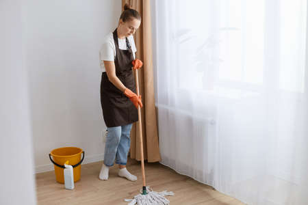 Indoor shot of dark haired female with hair bun wearing white t shirt, apron and jeans cleaning her flat, washing floor with mop, doing household chores.の写真素材