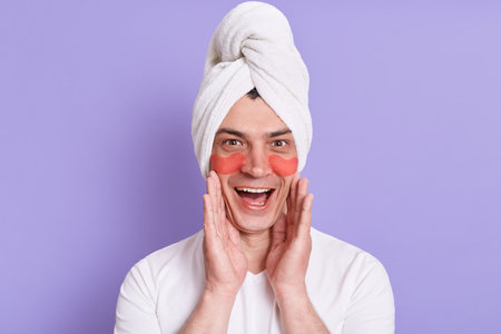 Funny portrait of man wearing bath towel on head, posing with glue sticky hydrogel eye-patches, doing cosmetic procedures, looking at camera with excited expression and raised arms.の写真素材