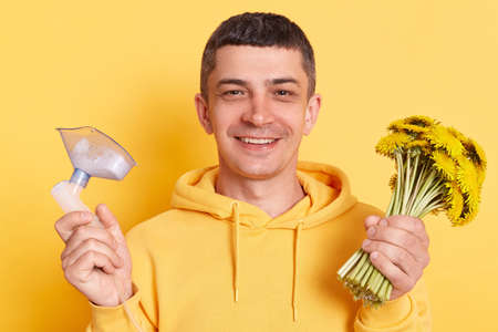 Smiling happy man wearing casual style hoodie posing isolated over yellow background, holding bouquet of dandelions and inhaler mask, looking at camera.の写真素材