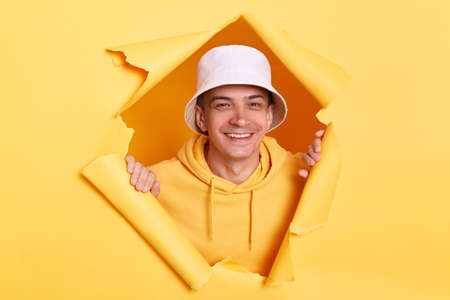 Portrait of positive joyful man wearing casual hoodie and panama looking through torn hole in yellow paper, looking at camera with toothy smile, expressing happiness.の写真素材
