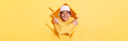 Portrait of smiling satisfied man wearing casual hoodie and panama looking through torn hole in...