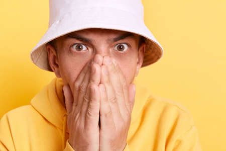Closeup portrait of shocked young adult man wearing casual style hoodie and panama, looking at camera with big eyes, covering face with palms, posing isolated over yellow background.の写真素材
