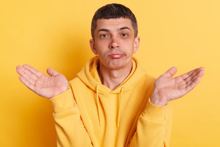 Man wearing casual style hoodie, posing isolated over yellow background, gesturing i don't know,...