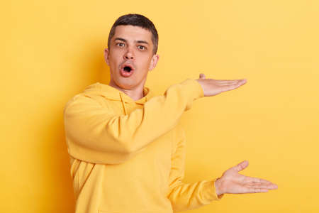Portrait of amazed man wearing casual style hoodie posing isolated over yellow background, looking at camera with open mouth and big eyes, showing huge size.の写真素材