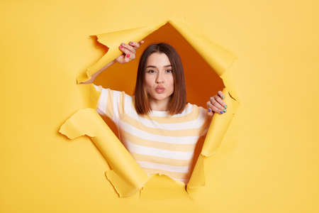 Portrait of romantic lovely adorable woman stands in torn paper hole, posing with pout lips and...