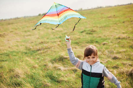 Portrsait of happy little girl flying a colorful kite running and jumping on a green meadow, wearing coat, spending day in active way, having fun alone, running happily.の写真素材