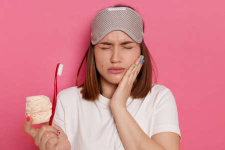 Indoor shot of sad depressed woman wearing white t shirt standing with sugary sweets and toothbrush in hands, feels pain in teeth, standing with closed eyes, keeping hand on cheek.の写真素材