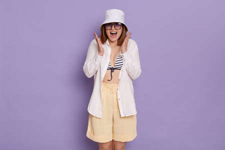 Portrait of delighted young Caucasian woman wearing swimsuit, shirt, shorts and panama posing isolated over purple background, being surprised, raised arms, screaming happily.の写真素材