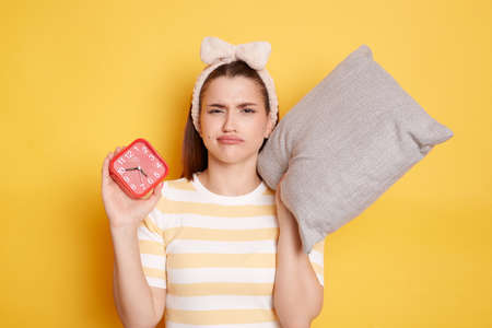 Indoor shot of sad woman with alarm clock awakening hard early wearing casual striped t-shirt posing...