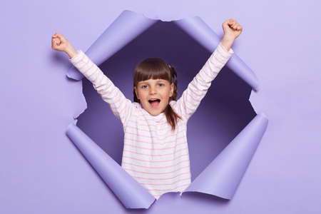 Portrait of amazed excited little girl with braids wearing striped shirt posing in torn paper wall, kid standing with raised arms, screaming happily, celebrating her success.の写真素材