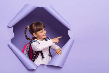 Horizontal shot of dark haired little girl with braids wearing striped shirt posing in torn paper wall, standing with backpack and pointing at copy space for advertisement.の写真素材