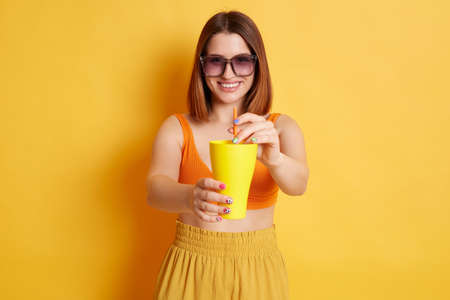 Portrait of smiling kind Caucasian woman in sunglasses wearing summer clothing posing isolated over yellow background, offering fresh beverage to you, holding cup with fresh delicious drink.の写真素材