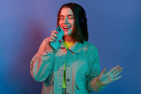 Image of Caucasian dark haired woman wearing stylish jacket standing with closed eyes, listening music, holding cell phone as a microphone and singing, posing isolated on neon light background.の写真素材