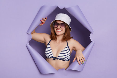 Indoor shot of smiling satisfied woman wearing striped swimming suit posing in torn purple paper wall, looking at camera with positive facial expression, being happy.の写真素材