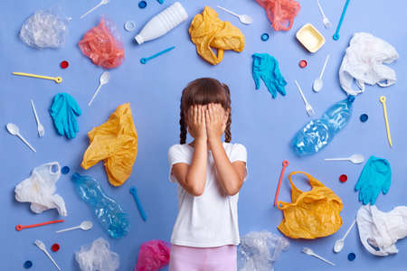 Horizontal shot of scared little girl in white T-shirt, picked plastic litter isolated on blue wall, covering eyes with palms, hiding her face, posing surrounded with much garbage.の写真素材