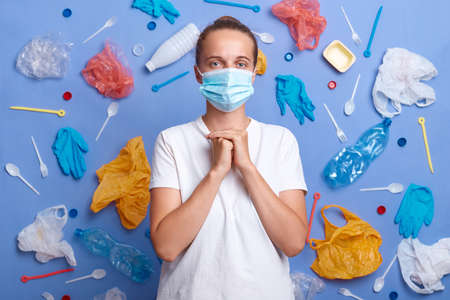 Caucasian woman wearing protective mask and white T-shirt standing against blue wall with symbolic much garbage, thinks how to solve problem of environmental pollution, keeps fists together.の写真素材