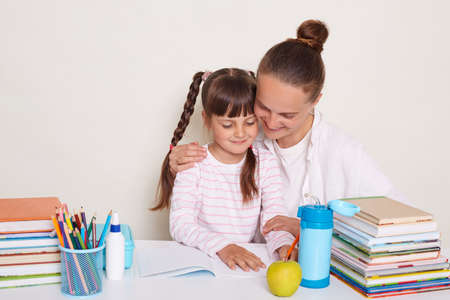 Indoor shot of young woman with bun hairstyle doing homework with daughter, family reading together, mother hugging her smart little schoolgirl, posing isolated over white background.の写真素材