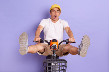 Full length of funny Caucasian delivery man in yellow cap riding bicycle, posing isolated over bright purple background. Fast and quick shipment service.の写真素材