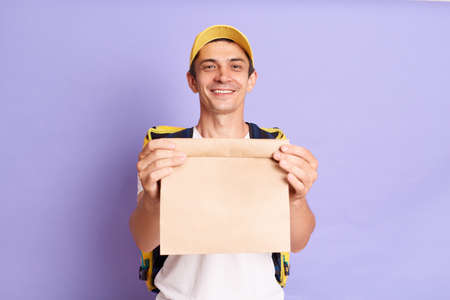 Satisfied delivery man in white t shirt and cap holding paper packet with food isolated over purple...
