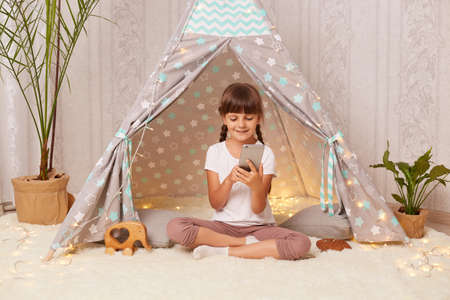 Horizontal shot of little girl sitting at home in teepee tent and playing on the phone, typing message or choosing cartoon for watching, sitting with crossed legs on floor and smiling.の写真素材