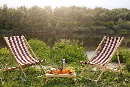 Outdoor shot of beautiful place for picnic, wooden table with food and arm chair for relaxing near...