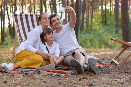 Image of happy positive satisfied family, husband, wife and their daughter sitting on the ground on blanket and taking selfie from their vacation in the forest, smiling happily.の写真素材