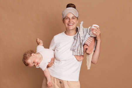 Horizontal shot of beautiful young woman wearing white t shirt and sleeping mask with baby in hands, standing looking at camera with charming smile, enjoying morning coffee.の写真素材