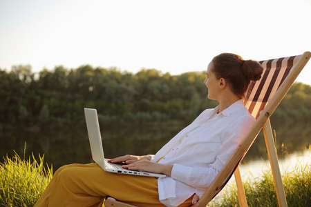 Side view of cam relaxed woman with bun hairstyle wearing white shirt sitting by the water in folding chair and working on laptop, looking away and enjoying beautiful nature.の写真素材
