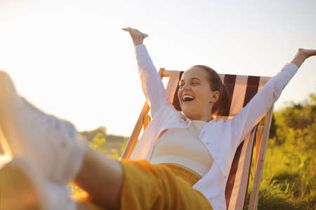 Horizontal shot of extremely happy dark haired Caucasian woman wearing white shirt sitting in the folding chair by the water, raised arms, looking at beautiful views, screaming with happiness.の写真素材