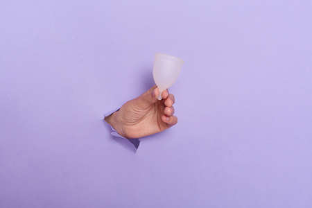 Photo of human's hand breaking out of a hole from a lilac background holding a menstrual cup for woman period, menstruation, healthcare.の写真素材