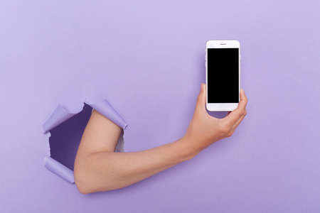 Indoor shot of person's hand holding phone from purple torn paper, smart phone with black blank screen for advertisement or promotional text.の写真素材