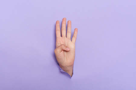 Person's hand sticks out of hole in paper of lilac background four fingers sign, copy space for advertisement.の写真素材