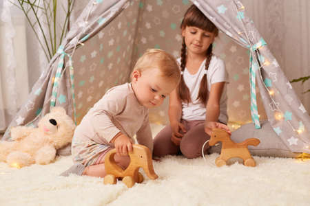 Horizontal shot of charming sibling girls playing together in peetee tent with wooden eco toys, toddler baby and her elder sister spending time together at home.の写真素材