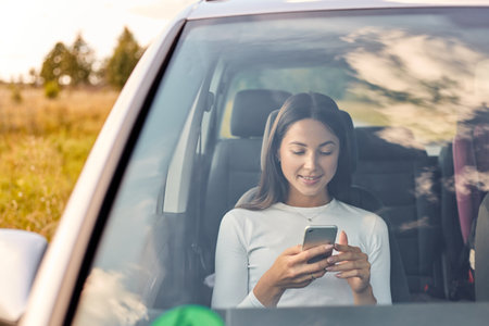 Smiling beautiful young adult woman looking at her mobile phone while sitting inside car, attractive...