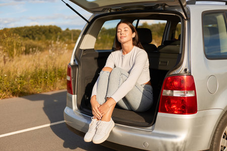 Portrait of young adult woman wearing white shirt and jeans sitting in car trunk, enjoying her travel, relaxing, enjoys sunshine, posing with closed eyes.の写真素材
