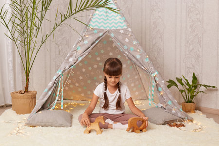 Portrait of dark haired cute little girl playing with toys and in teepee tent, sitting on floor on soft carpet, posing alone in wigwam, waiting with her friends.の写真素材