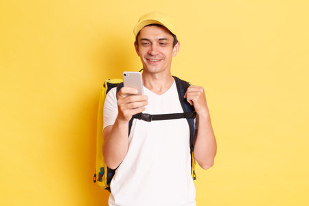 Attractive Caucasian delivery man in yellow cap and t-shirt with thermal backpack holding cell phone, looking at display, chatting while has free time, isolated on yellow background.の写真素材