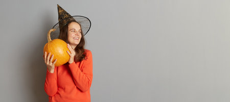 Indoor shot of smiling satisfied woman holding big ripe orange pumpkin, wearing sweater and witch...