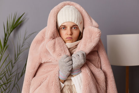 Indoor shot of sad upset Caucasian woman wearing cap, gloves, scarf and coat sitting in living room, puts on warm clothing while being at home in her cold apartment.の写真素材