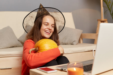 Indoor shot of smiling attractive female in witch hat sitting on floor and embracing orange pumpkin...