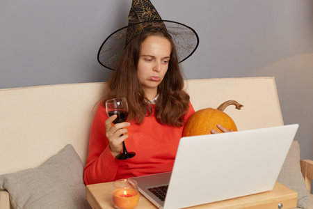 Horizontal shot of sad Caucasian woman wearing witch hat working on laptop in home interior and...