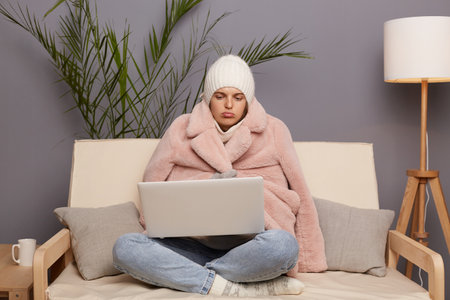 Horizontal shot of sad Caucasian woman posing in cold room sitting on the sofa wearing warm hat, mittens and cap, holding laptop on knees, looking at display with pout lips.の写真素材
