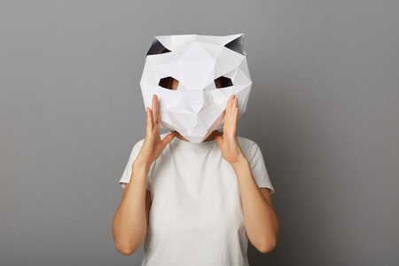 Horizontal shot of unknown female wearing cat mask and white t-shirt isolated over gray background, screaming loud, keeps hand near her mouth, making announcement.の写真素材