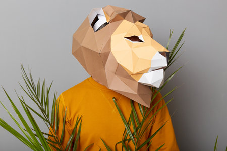 Profile portrait of unknown man wearing lion mask and orange jumper standing among green plant leaves looking away, posing isolated over gray background.の写真素材