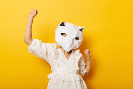Photo of overjoyed anonymous woman wearing dress and paper cat mask, raised her fists, celebrating her success and victory, posing isolated over yellow background.の写真素材