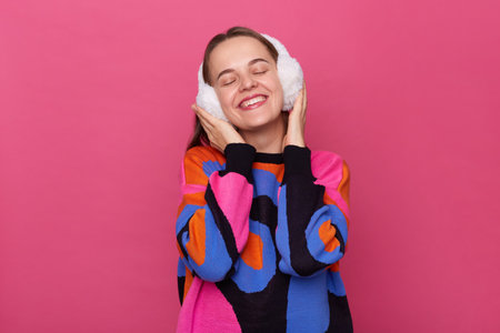 Portrait of overjoyed satisfied delighted woman wearing winter fluffy earmuffs and warm sweater, keeps hands on her head, standing with closed eyes and toothy smile isolated over pink background.の写真素材